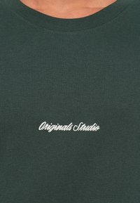 Tricou din bumbac de culoare verde închis, cu guler rotund, textură moale și textul brodat alb "Originals Studio" în zona pieptului.