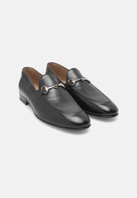 Mocassins en cuir noir avec un bout pointu, dotés d'une chaîne en métal doré sur le devant, et d'une finition lisse et polie.