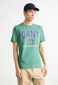 Zelené bavlněné tričko s velkým námořnickým textem "GANT" a logem, s textem "AMERICAN SPORTSWEAR" pod ním. Nosič má námořnickou čepici.