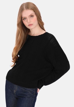 Fiatal nő hosszú, világosbarna hajjal, fekete kötött pulóvert és kék farmert visel, zsebébe tett kézzel pózol fehér háttér előtt.