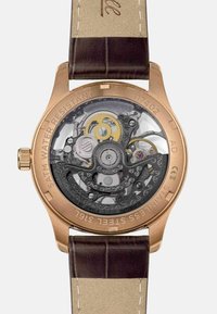 Montre automatique avec un fond de boîte transparent. Caractéristiques : boîte en bronze, bracelet en cuir brun, engrenages complexes et 22 pierres précieuses. Résistante à l'eau jusqu'à 5 ATM.
