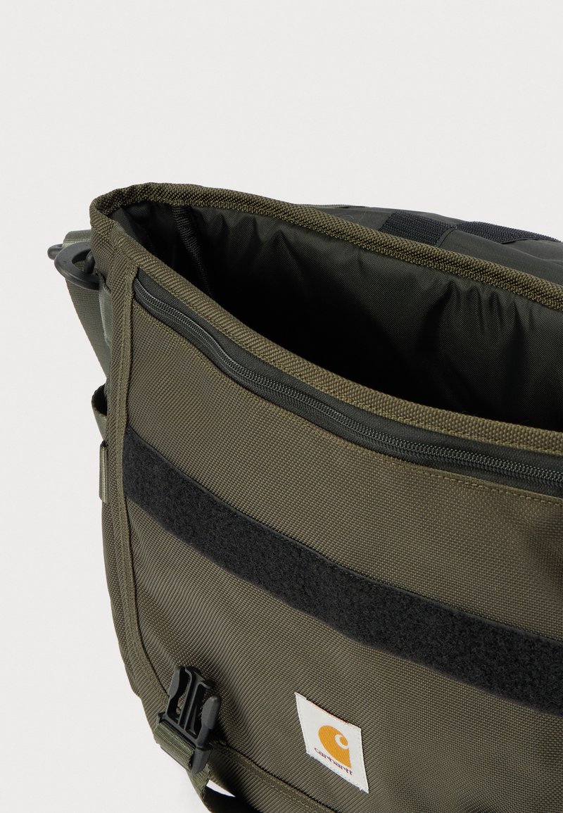 Sac d'épaule en tissu vert avec fermeture éclair noire, bande velcro, boucle en plastique et écusson logo Carhartt sur la poche avant.