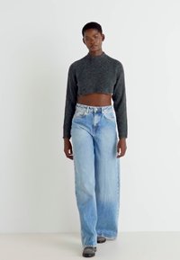 Pulover scurt, de culoare gri închis, cu guler rotund; blugi deschiși la culoare, cu croială largă și un model decolorat; model purtând încălțăminte neagră.