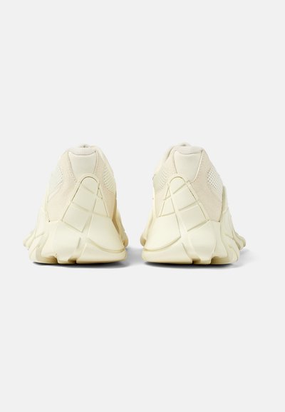 Cream-colored sneakers with textured mesh and suede panels, featuring a rubber sole and angular design. Seam detail and padded collars.