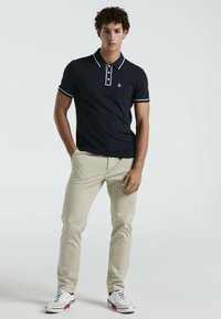 Polo marine à bordure blanche avec patte de boutonnage à trois boutons, associé à un pantalon beigne et des baskets blanches avec des accents rouges et bleus.