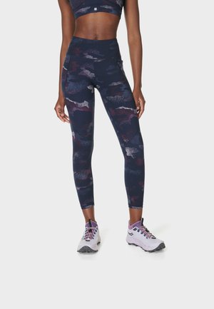 Vrouw die marineblauwe en paars camouflerende legging met zijzakken en lichtpaarse trailrunningschoenen draagt.