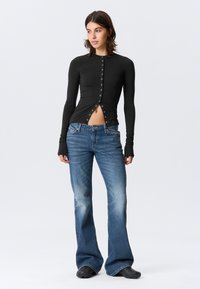 Haut noir à manches longues côtelé avec boutons sur le devant et fentes, associé à un jean évasé bleu délavé. Tissu texturé et silhouette ajustée.