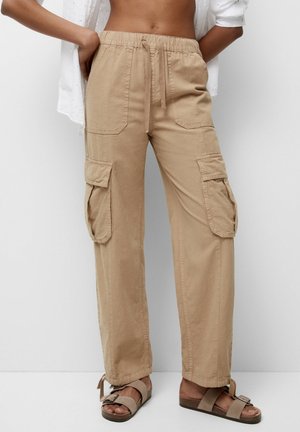 Beige cargobroek met meerdere zakken, een taille met trekkoord, gedragen met lichtbruine sandalen met gesp en een gedeeltelijk zichtbaar wit overhemd.