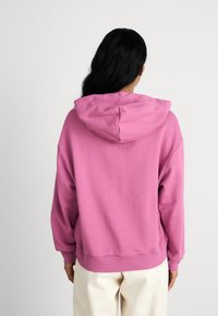 Pink hooded sweatshirt with a relaxed fit, featuring a kangaroo pocket and ribbed cuffs. Worn over cream-colored pants.