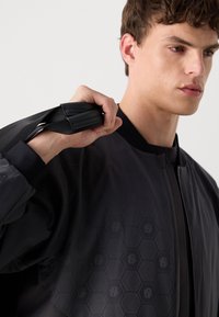 Black bomber jacket with hexagonal texture, ribbed cuffs, and a straight zipper. Fabric appears smooth and lightweight, featuring subtle branding.