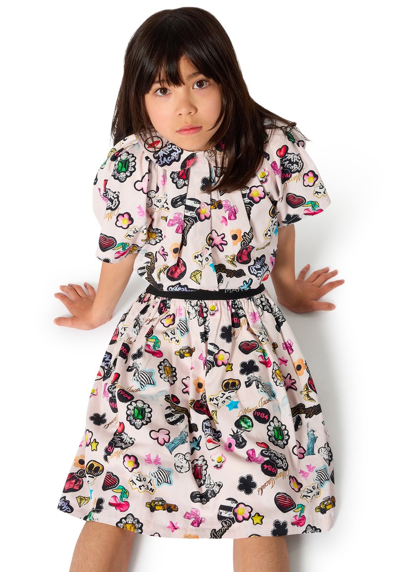 Junges Mädchen mit dunklem Haar, das ein bunt gemustertes Kleid trägt und vor weißem Hintergrund sitzt, schaut direkt nach vorne.