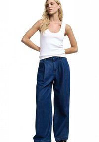 Canottiera bianca abbinata a pantaloni in denim scuri, larghi e a vita alta, con pieghe. L'outfit presenta un taglio casual e una texture morbida.