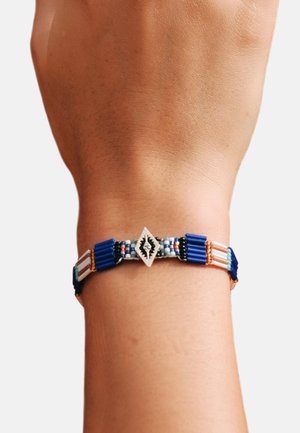 Bracelet en perles présentant des matériaux bleus, blancs et noirs, avec un charme central en forme de diamant et des motifs de perles complexes.
