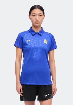 Młoda kobieta ubrana w niebieską koszulkę piłkarską Chelsea z żółtymi logo i czarne spodenki Nike, stoi na jednolitym tle.