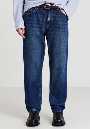Jeans di denim blu con taglio ampio, caratterizzati da cinque tasche, passanti per cintura e una texture liscia. Indossati con una cintura marrone e stivali neri.