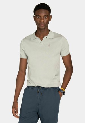 Uomo che indossa una polo a maniche corte grigio chiaro e pantaloni con coulisse blu scuro, in piedi con una mano in tasca davanti a uno sfondo semplice.