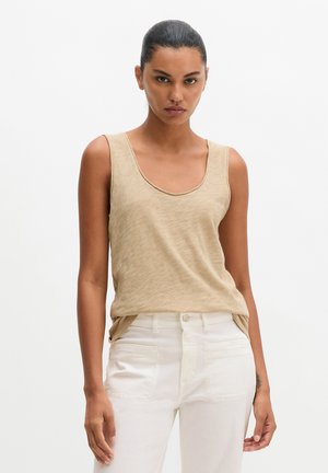 Femme aux cheveux foncés attachés derrière la tête, portant un top beige sans manches et un pantalon blanc taille haute, debout devant un fond clair uni.