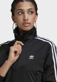 Mujer vestida con una chaqueta de chándal negra de Adidas con rayas blancas, sosteniendo la cremallera cerca del cuello, mirando hacia un lado, lleva aretes grandes en forma de aro.