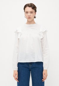 Kvinne iført en hvit langermet gjennomsiktig bluse med volanger og blonde detaljer, parret med blå jeans, stående mot en ensfarget bakgrunn.