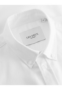 Camicia bianca con colletto classico, caratterizzata da una texture liscia, etichetta cucita e due chiusure a bottone sul colletto.