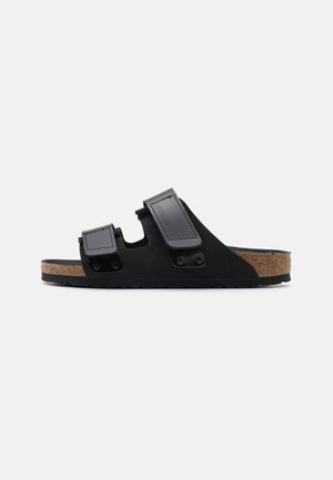 Sort slidesandal med to justerbare remme og korkfodsål på hvid baggrund.