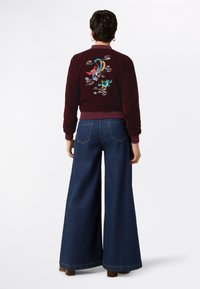 Giacca bomber bordeaux con motivo ricamato colorato sulla schiena, abbinata a jeans in denim blu a gamba larga. Tessuto morbido.