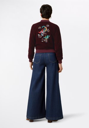 Bordeauxfarbene Bomberjacke mit buntem besticktem Design auf dem Rücken, kombiniert mit weiten blauen Jeans. Glatte Stofftextur.