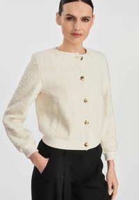 Biała, strukturalna kurtka z okrągłym dekoltem, złotymi guzikami i prążkowanymi mankietami. Połączona z czarnymi spodniami o luźnym kroju.