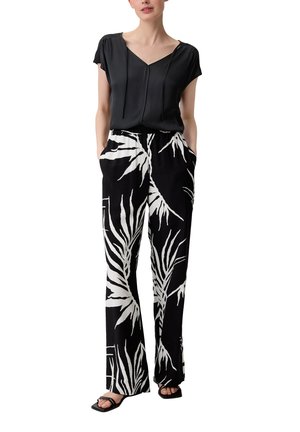 Kvinde iført sort kortærmet bluse og sorte bukser med vidde og store hvide bladmønstre, stående med hænderne i lommerne.