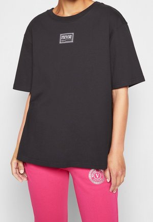 Osoba ubrana w czarny T-shirt Versace Jeans Couture i różowe spodnie z logo Versace na lewym udzie, stojąca na jednolitym tle.