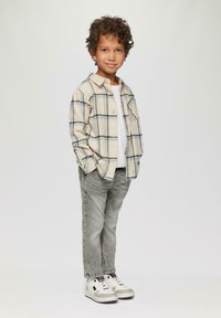 Geruit beige overhemd met blauwe accenten, grijze jeans en witte sneakers. Kind staat met handen in de zakken en glimlacht.