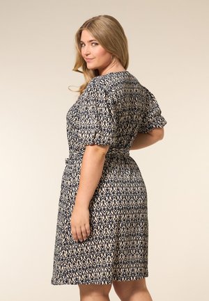 Een vrouw met golvend blond haar poseert in profiel, gekleed in een gedessineerde jurk met pofmouwen, en glimlacht zelfverzekerd naar de camera.