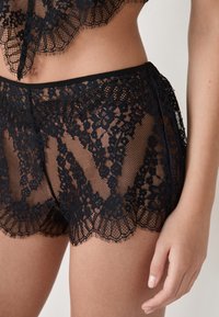 Conjunto de lencería de encaje negro que incluye tejido transparente, patrones florales, bordes en puntilla y un diseño ajustado. Incluye pantalones cortos y una parte superior a juego.