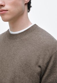 Brązowy sweter z dzianiny z okrągłym dekoltem i prążkowanym kołnierzem. Miękka faktura z delikatnymi różnicami w kolorze, widoczne subtelne detale.