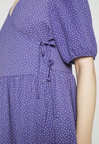 Robe violette à pois blancs ; dotée de manches courtes bouffantes, d'une taille cintrée avec des liens, et d'une jupe fluide. Tissu doux et texturé.