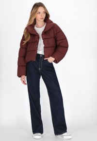 Giacca a vento marrone con colletto alto, chiusura a bottoni e texture trapuntata, abbinata a jeans in denim scuro a gamba larga e sneakers bianche.