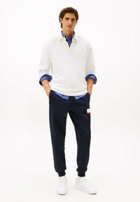 Weißer Pullover mit Kragen, über einem blau-weiß gestreiften Hemd getragen; dunkelblaue Jogginghosen mit rotem und weißem Akzent; weiße Sneakers.