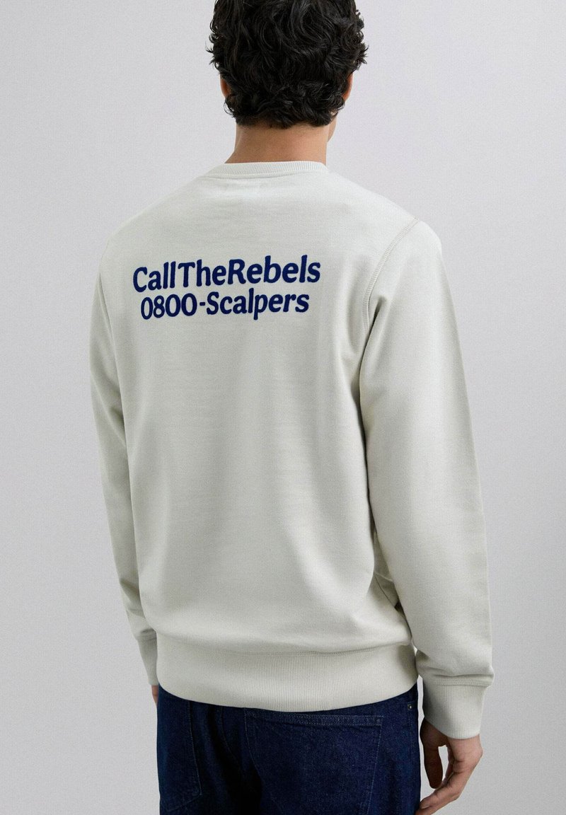 Cremefarvet sweatshirt med blå broderet tekst "CallTheRebels" og "0800-Scalpers" på ryggen. Ribbet kant ved ærmer og bund.