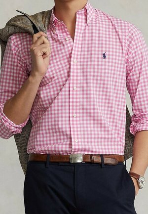 Homme portant une chemise à carreaux rose et blanche boutonnée, pantalon sombre, ceinture marron, tenant une veste sur l'épaule avec les manches retroussées.