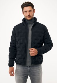 Černá péřová bunda s předním zipem, úzkými manžetami a vysokým límcem, s prošívaným designem a dvěma postranními kapsami. Nositá přes šedý svetr.