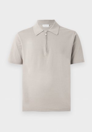 Homme debout, les mains dans les poches, portant un polo beige à manches courtes avec fermeture éclair, un pantalon marron et des chaussures habillées marron foncé sur fond blanc.