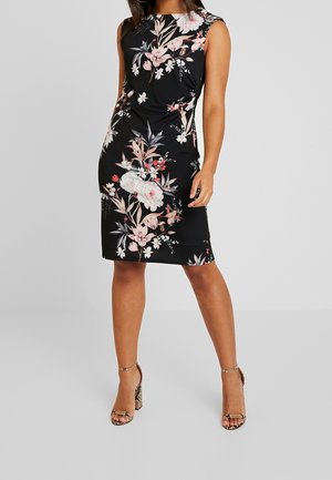 Frau trägt ein ärmelloses schwarzes knielanges Kleid mit rosa und weißen Blumenmustern und hohe offene Sandalen mit Absatz.