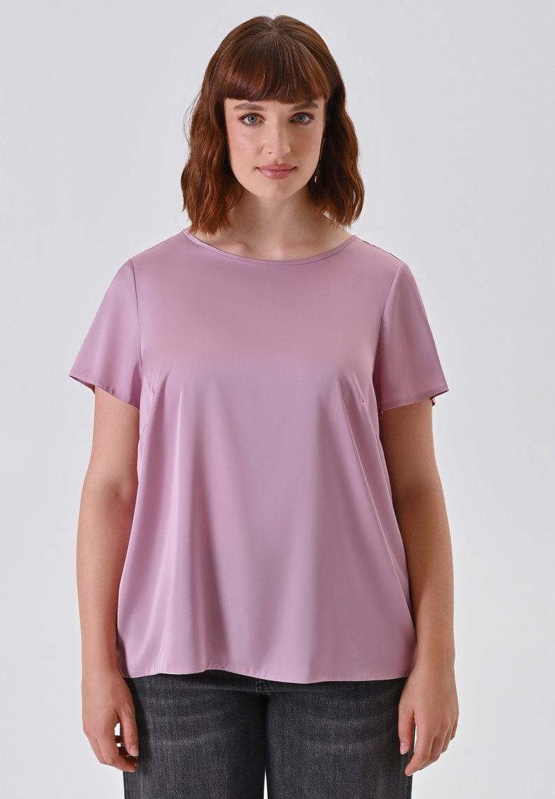 Frau mit schulterlangem braunem Haar, die eine lockere rosa kurzärmlige Bluse und dunkle Jeans trägt, steht vor einem schlichten hellen Hintergrund.