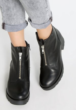 Bottes cheville en cuir noir avec fermetures éclair argentées à l'avant, portées avec un jean gris clair retroussé sur fond blanc.