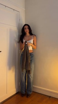 Bílý krátký tílko s krajkovým lemem, modré volné džíny a hnědé kozačky na vysokém podpatku. Drží tmavě pletený šátek. Neutrální pozadí.