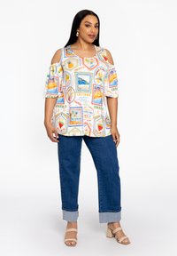 Kurzarmbluse mit einem bunten Reisethema-Print, weißem Hintergrund und Cold-Shoulder-Design, kombiniert mit hochgekrempelten blauen Jeans und beigen Sandalen.