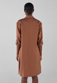 Brązowa, długoshirtowa sukienka z długim rękawem, zapinana na guziki, wykonana z gładkiego materiału. Posiada podwinięte rękawy, boczne rozcięcia i luźny krój dla wygody.