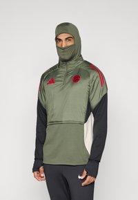 Αθλητικό μπουφάν σε ελιά πράσινο με κουκούλα και κάλυμμα προσώπου, με κόκκινες ρίγες και λογότυπο. Κατασκευασμένο από απαλό ύφασμα με σχεδίαση σε στρώσεις.