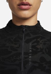 Černý top s polovičním zipem, který má texturovaný vzor, šedé logo "NIKE TRAIL", stojáček a atletický střih.
