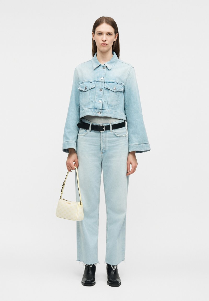 Femme portant une veste en jean courte bleu clair et un jean, ceinture noire, bottes noires, tenant un petit sac à main crème, debout devant un fond uni.